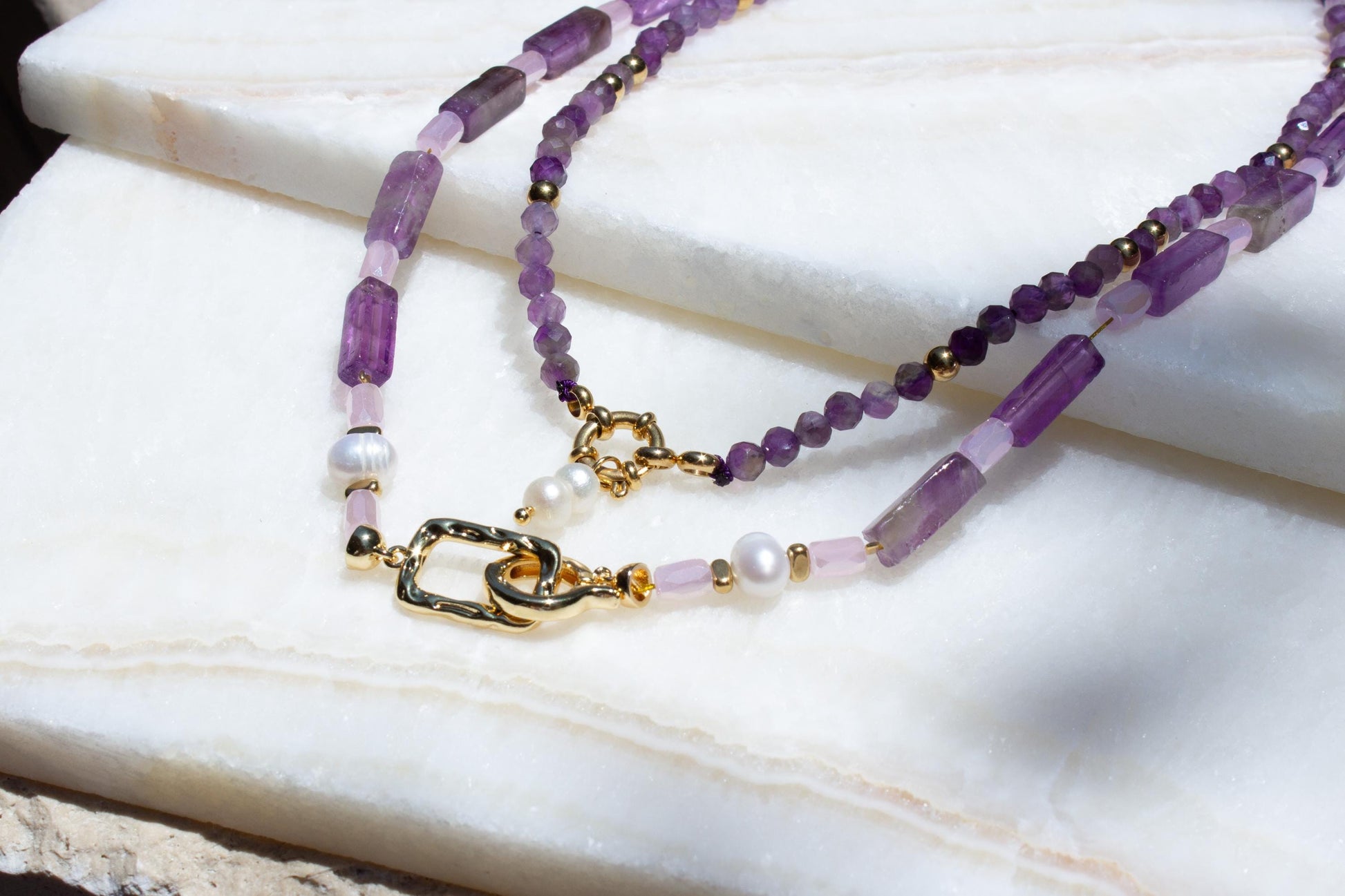 two amethyst necklaces layered on two white tiles.