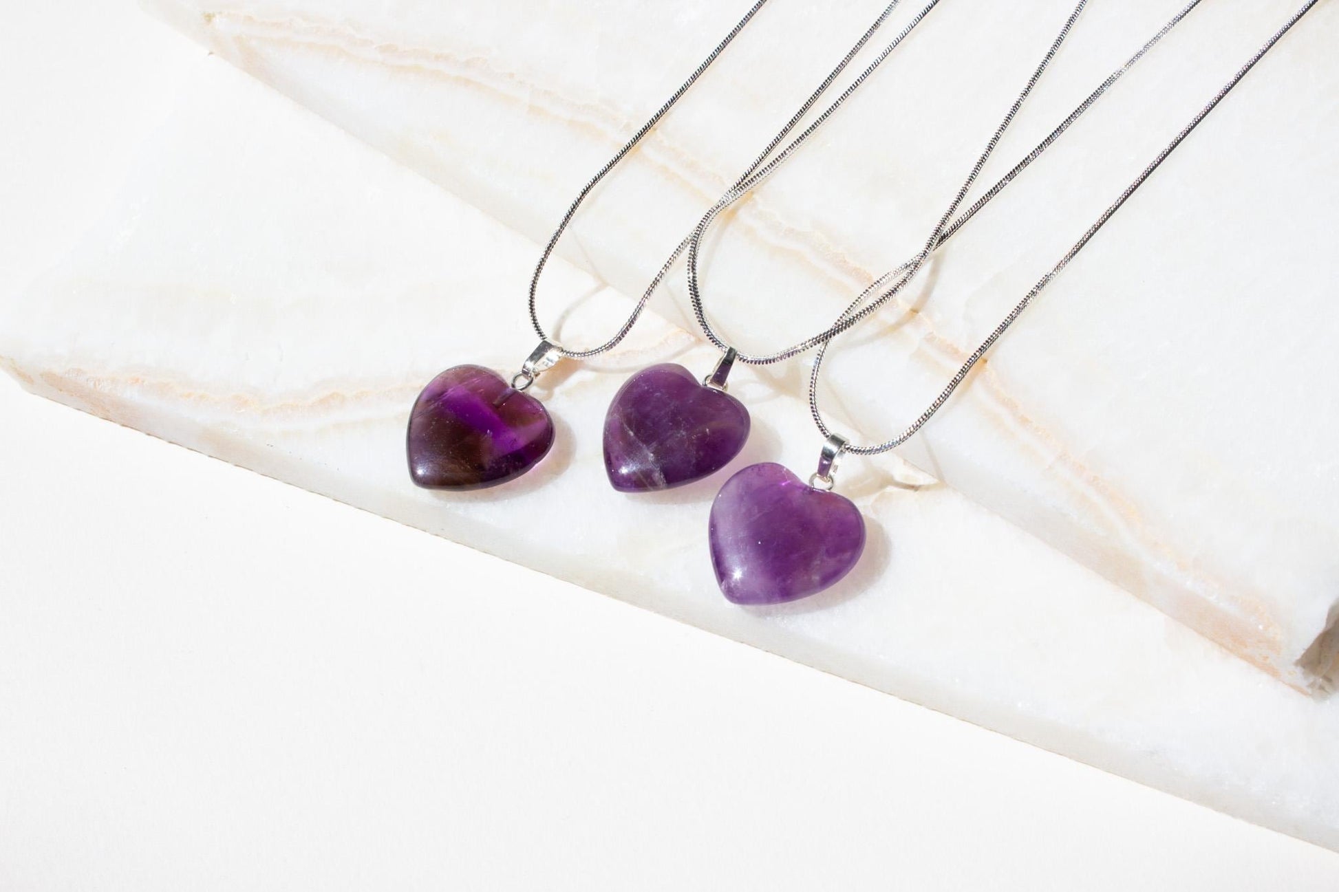three amethyst heart necklaces laying on white tiles.