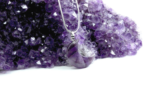 one purple crystal moon necklace leaning against a large amethyst crystal. Both in vibrant purple colors.