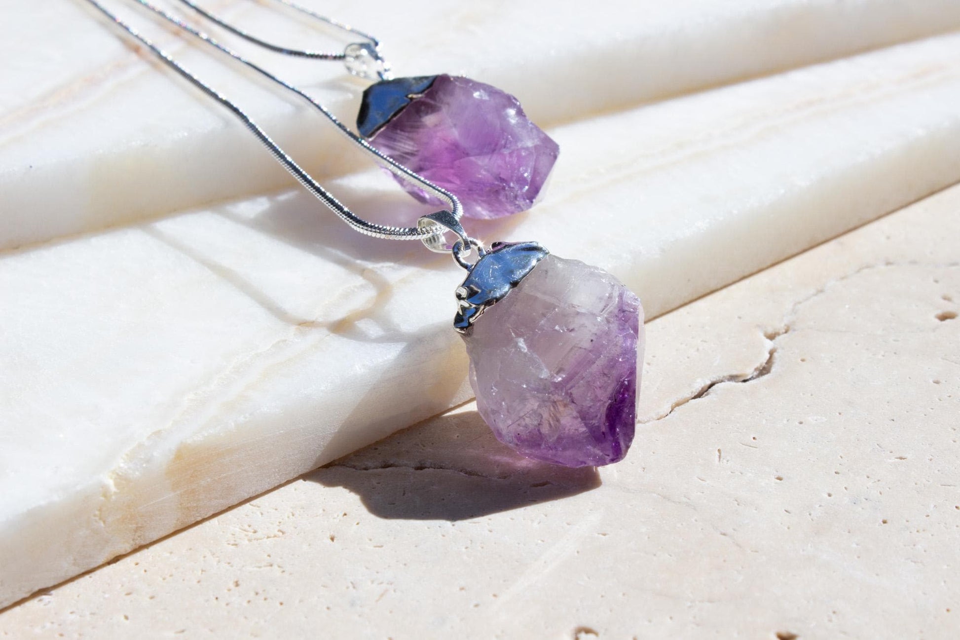 two amethyst raw stones on beige tile. Both pendants have silver tops.