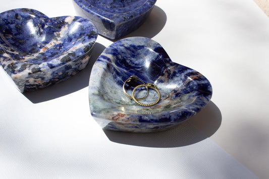 group of small heart-shaped dishes in a blue sodalite crystal gemstone. One of the dishes is holding two gold rings.