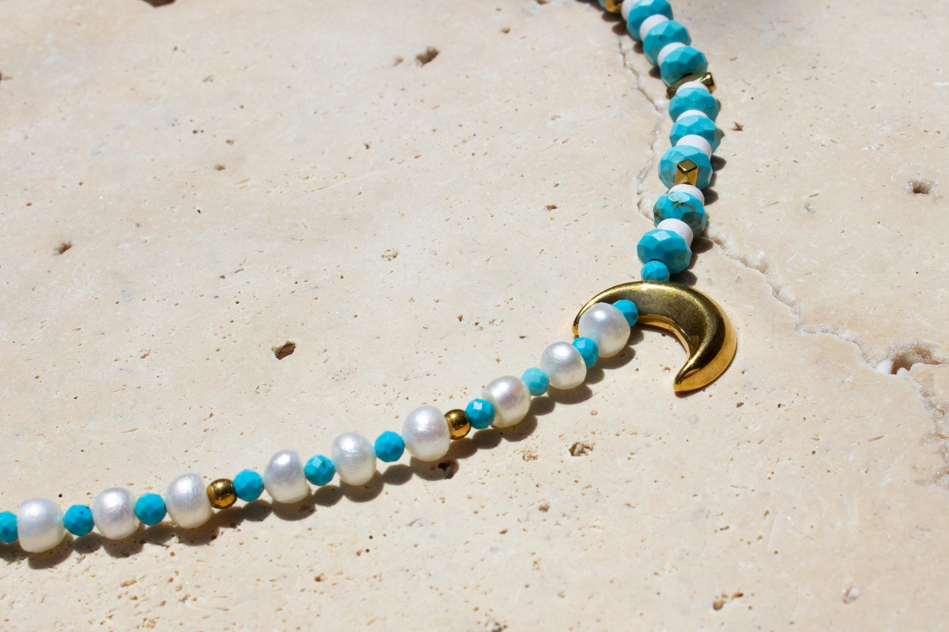 pearl and turquoise beaded choker on a concrete tile. There's a gold moon pendant in the middle.